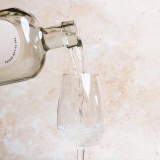 58 and CO Olive Oil Vodka Greek bottle pouring liquid into a glass on a light marble surface.