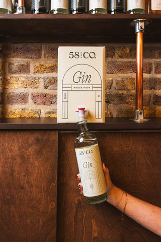 Person holding a bottle of 58 and CO Gin being refilled from a 5L BIB in front of a brick wall with a shelf displaying bottles.