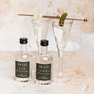 Two miniature bottles of 58&Co Gin Martini and Vodka Martini with two cocktails garnished with a pickle and a cherry on a marble surface.