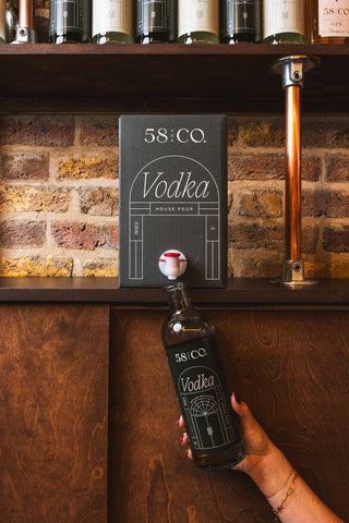 Person holding a bottle of 58 and CO Vodka being refilled from a 5L BIB in front of a bar setup with shelves and a brick wall.