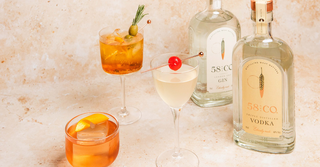 Three cocktails in glasses with garnishes next to two bottles of 58 & Co. vodka on a light stone surface.
