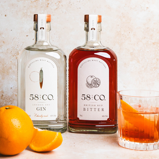 Two bottles of 58 Co Gin and Bitter with a glass of negroni cocktail and oranges on a light background