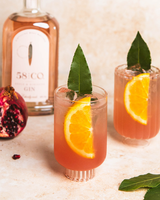 Two glasses of Pomegranate Bay Spritz cocktail with orange slices and a bottle of 58 and CO Apple & Hibiscus Gin on a light surface.