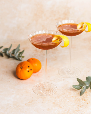 Two Clementine & Clove Chaplin cocktails with lemon twists and oranges on a light surface