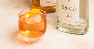 Negroni cocktail in a glass with a large ice cube and orange peel twist next to a bottle of 58 and CO gin on a light surface.