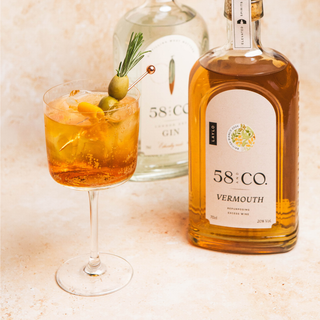 Bottle of 58 and Co vermouth with a Dirty Martini spritz cocktail garnished with olives and a rosemary sprig on a light background