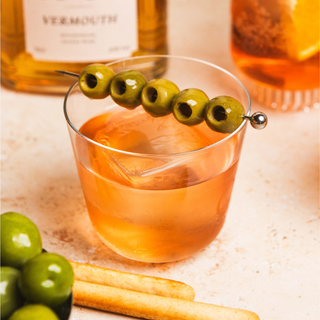 Dirty Negroni Cocktail in a glass, garnished with five olive on a metal pick, on a light surface with a bottle of vermouth in the background.