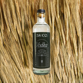 A clear glass bottle of 58 and CO House Pour Vodka with a dark green label and cork stopper, centered against a background of golden wheat stalks.