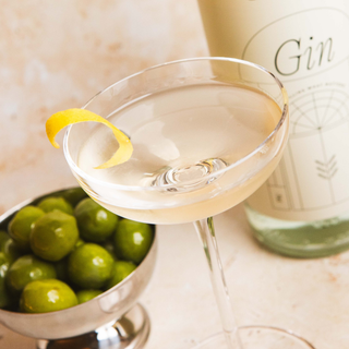 Martini Cocktail glass with lemon twist and olives next to a bottle of 58 and CO House Pour Gin on a marble surface.