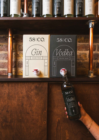 Bottle of 58 and CO Vodka held by a hand in front of a wooden shelf, being refilled from a 5L BIB