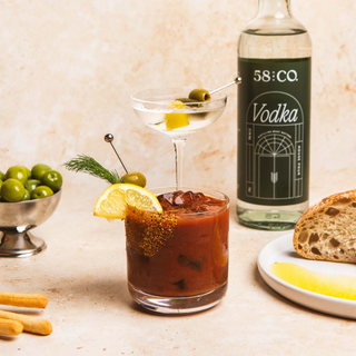Vodka martini and bloody mary cocktail with garnishes, bottle of 58 and CO House Pour Vodka, and bread on a table.