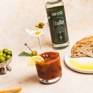 Bloody mary with lemon wedge and olives, 58 and CO House Pour vodka bottle, and bread on a light background