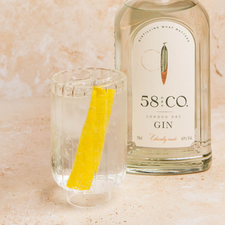 58 and CO London Dry Gin bottle with a glass of gin and tonic garnished with a lemon peel on a beige marble surface.
