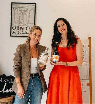 Carmen O'Neal and Sarah Vachon holding bottles of Olive Oil Vodka and extra virgin oil respectively.