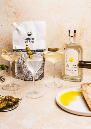 Citizens of Soil Olive Oil Pouch, a bottle of 58 and CO Olive Oil Vodka and martini cocktails with snacks and olive oil being poured onto a plate