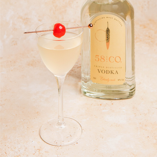 Bottle of 58 and Co triple distilled vodka with a pina colada martini cocktail garnished with a cherry on a light background