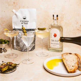 A styled tabletop scene with a bottle of 58 and CO olive oil vodka, a Citizens of Soil olive oil club pouch, three martini glasses, olives, bread, and a plate with olive oil on a beige stone surface.