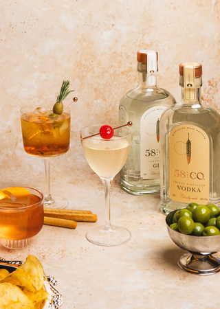 Two cocktails with garnishes, bottles of 58 and Co vodka and gin, and a bowl of olives on a textured surface.
