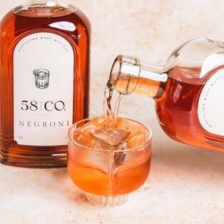 A bottle of 58 and CO NEGRONI cocktail and a glass of the amber-colored drink being poured into a rocks glass with ice, on a warm beige surface.