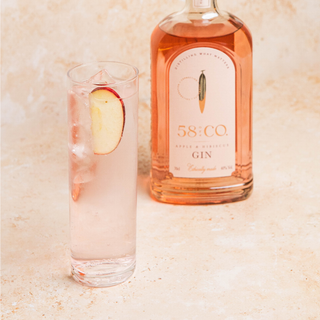 Bottle of 58 and CO Apple & Hibiscus Pink Gin with a glass of pink gin and tonic on a beige background