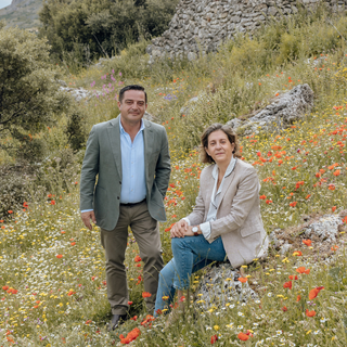 The Lopez Family - A man in a green blazer and a woman in a beige blazer posing together in a sunny, grassy hillside field filled with red and white wildflowers.